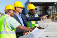 Three construction workers on a job site in hard hats and safety vests reviewing a blueprint.